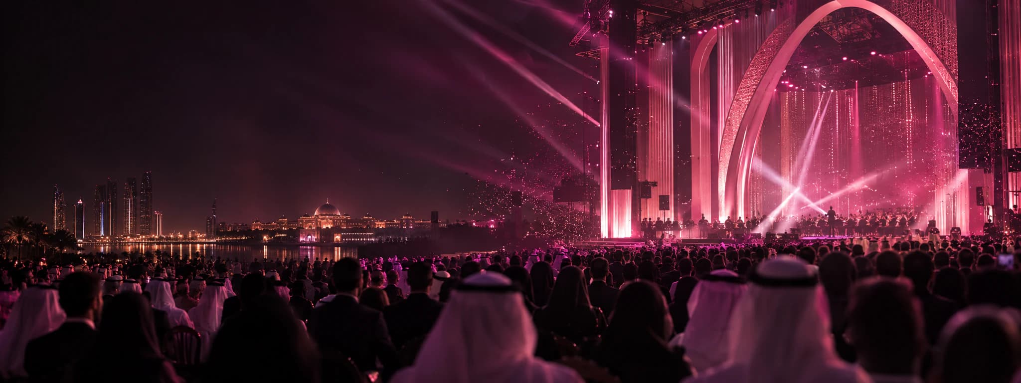 An evening concert on an outdoor stage lit in pink: an orchestra plays beneath a tall arch before a seated audience, with a city skyline across the water.