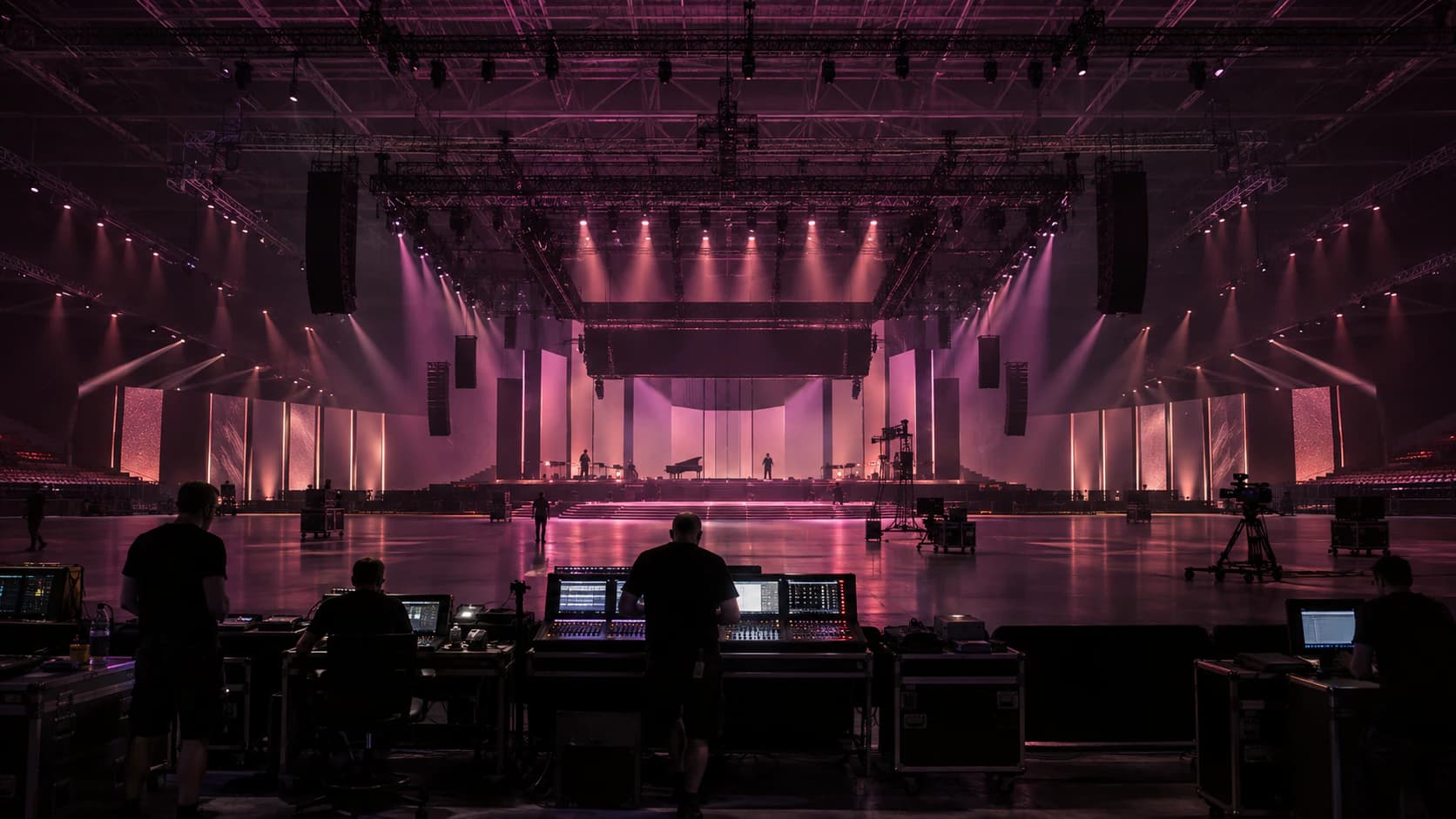 Technicians at sound and lighting consoles face a large, empty arena stage under lighting rigs glowing pink.