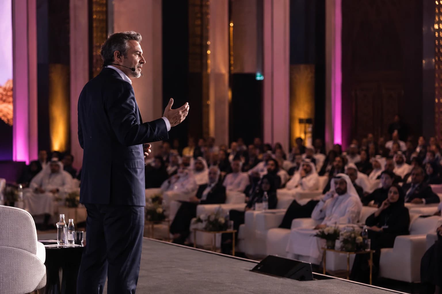 A speaker wearing a headset microphone addresses a seated audience from a stage.