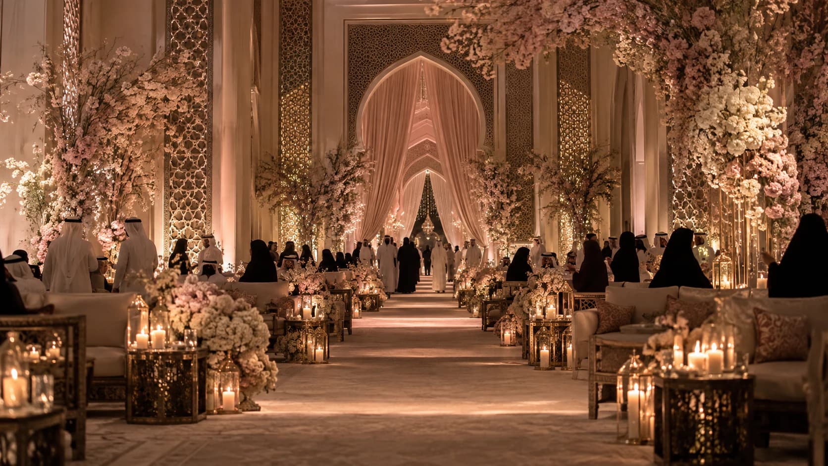 A long candlelit hall decorated with flowers, arches and lanterns, with guests seated on sofas along a central aisle.